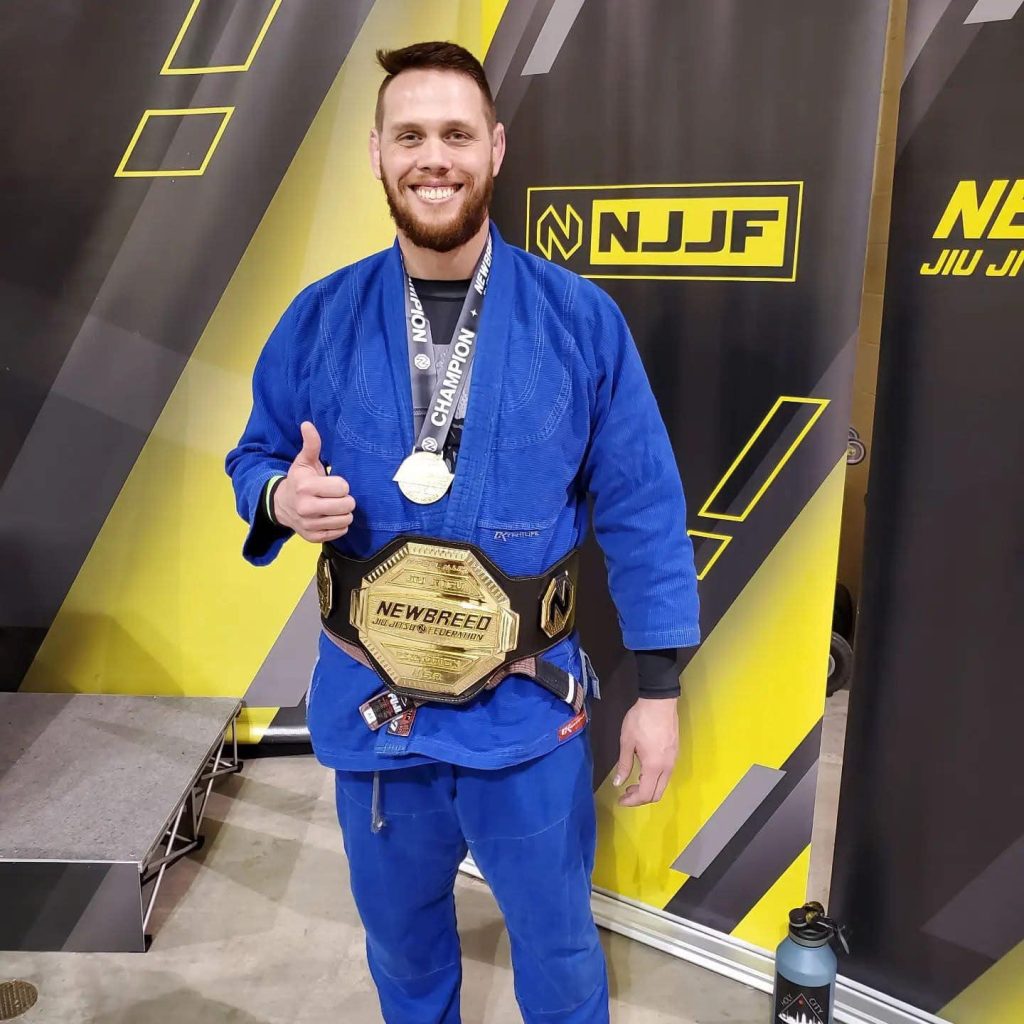 BJJ athlete wearing blue gi, holding championship belt, in front of NJJF logo, showcasing victory.