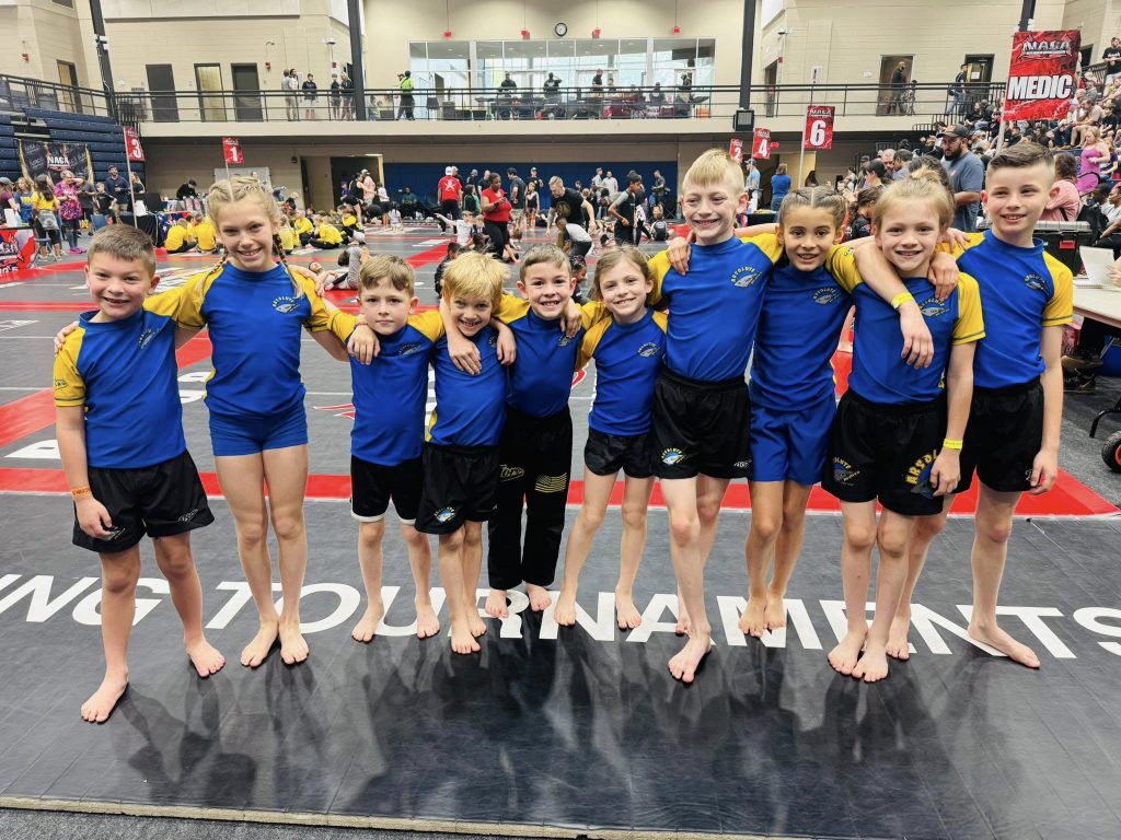 Kids standing together on competition mat with arms around each other, smiling cheerfully.