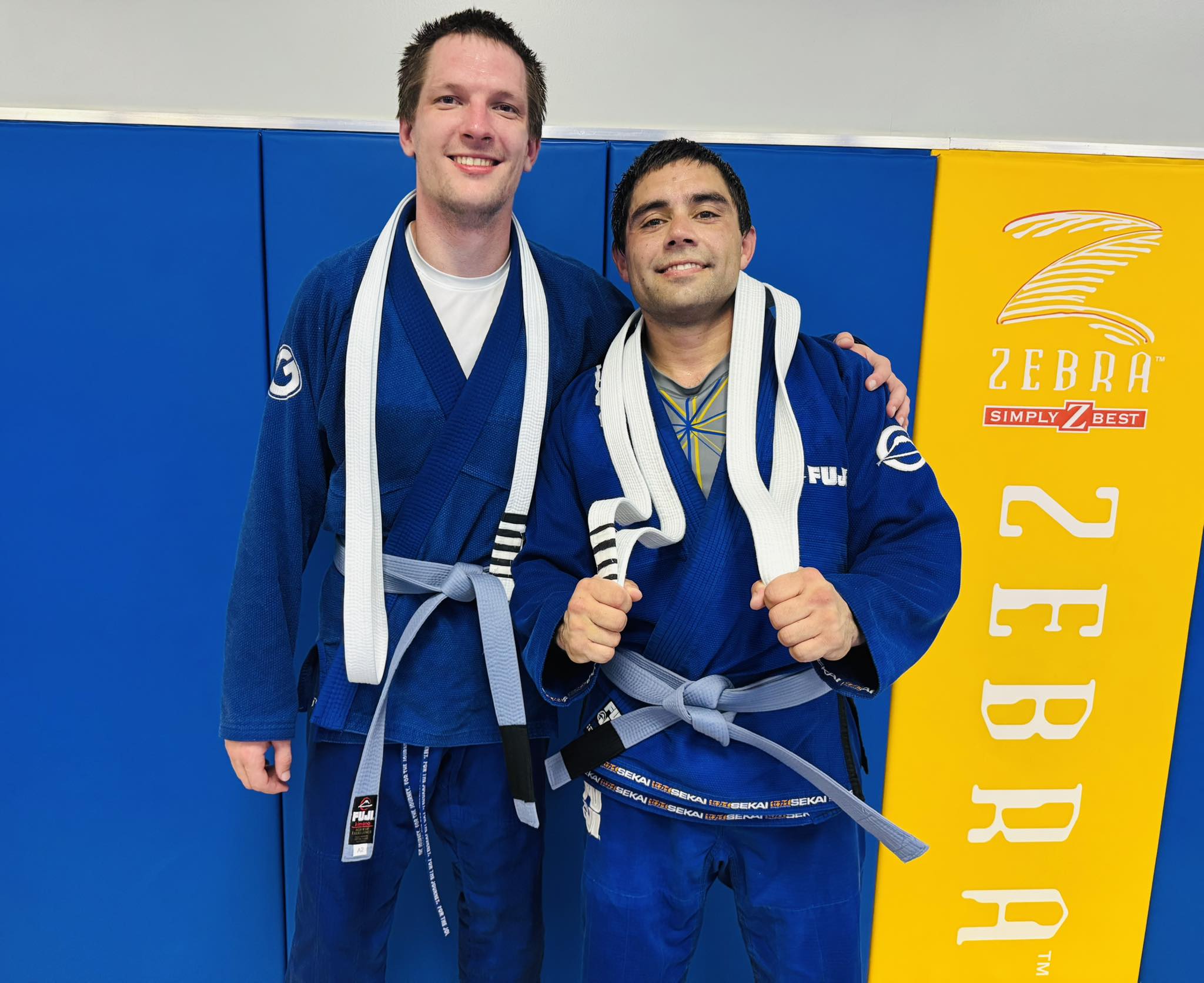 Two male Brazilian Jiu-Jitsu practitioners in blue gis with white belts on a competition mat.