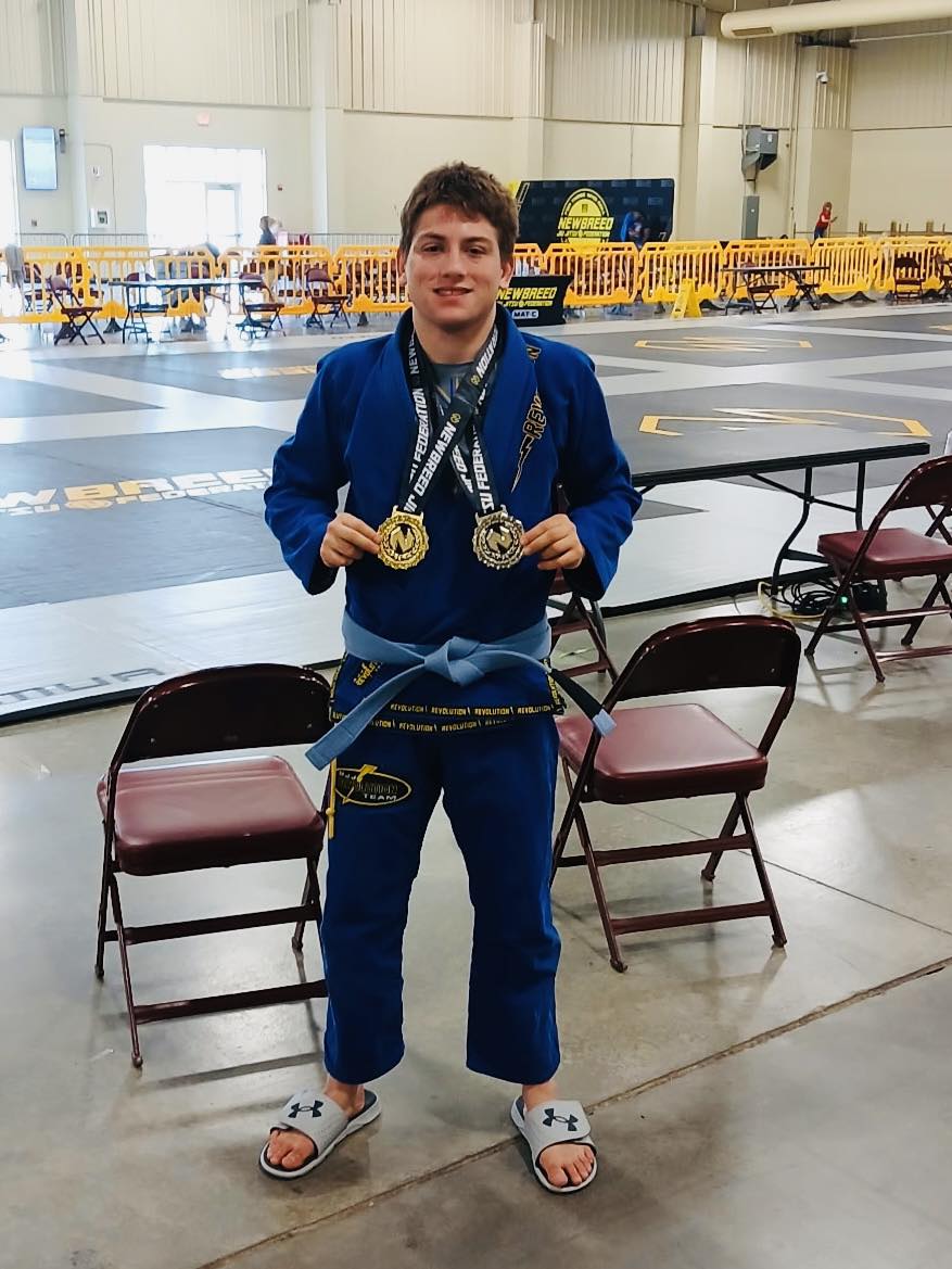 Young martial artist in blue gi with medals, celebrating victory at Brazilian Jiu-Jitsu tournament.