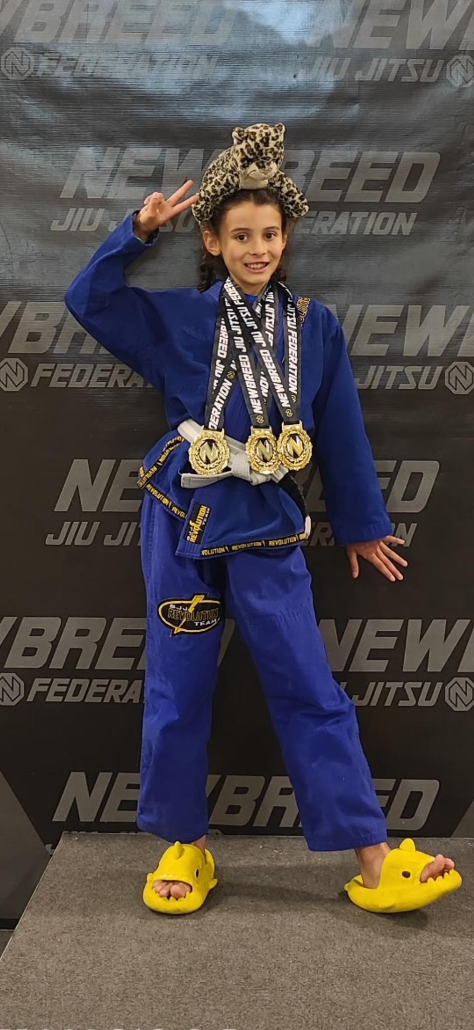 Young girl in a blue gi with medals, making victory signs at Absolute Jiu-Jitsu Academy.