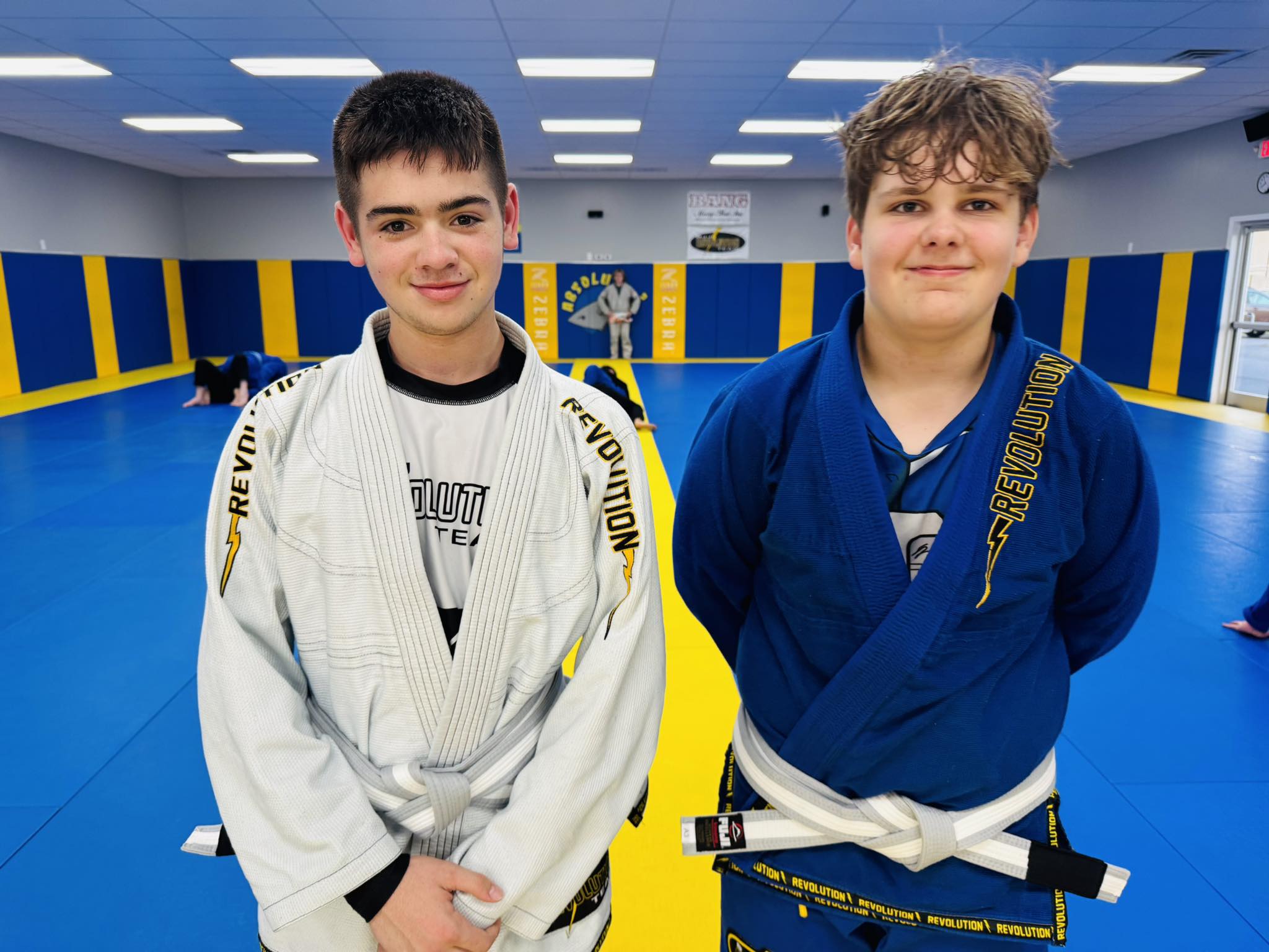 Alt text: Two young boys in Brazilian Jiu-Jitsu Gis smiling at the camera inside the academy.